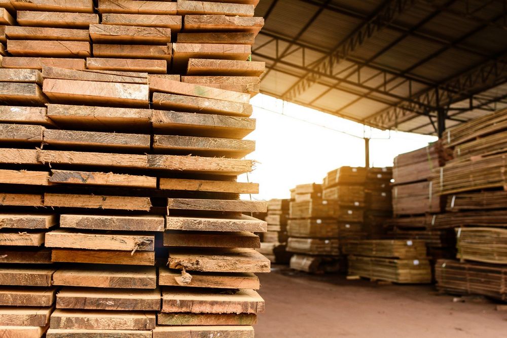 A pile of timber is shown with more piles in the background of a covered timber yard.