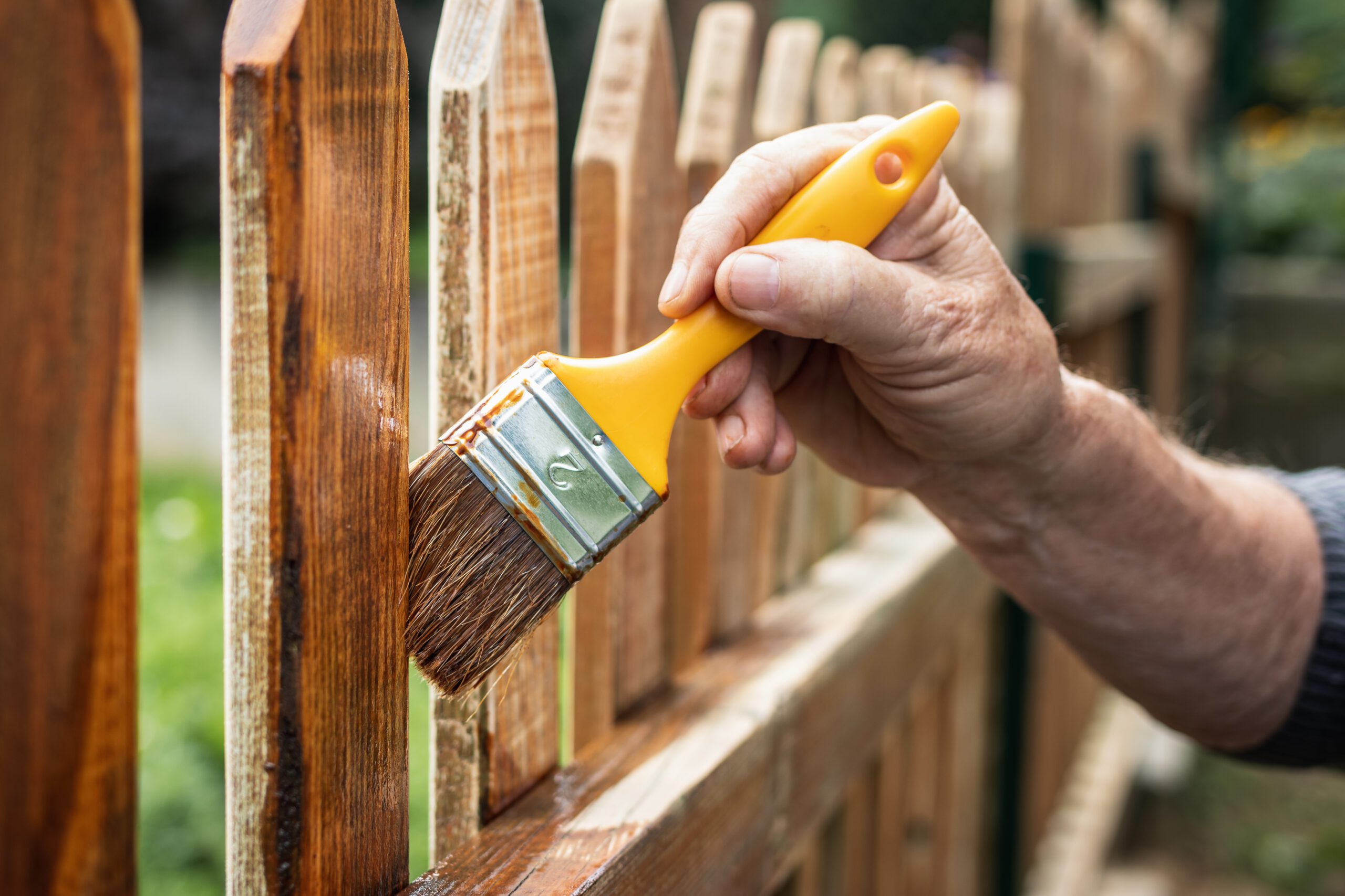 person using paintbrush to apply sealant to wooden fencing to waterproof the timber fence palings.