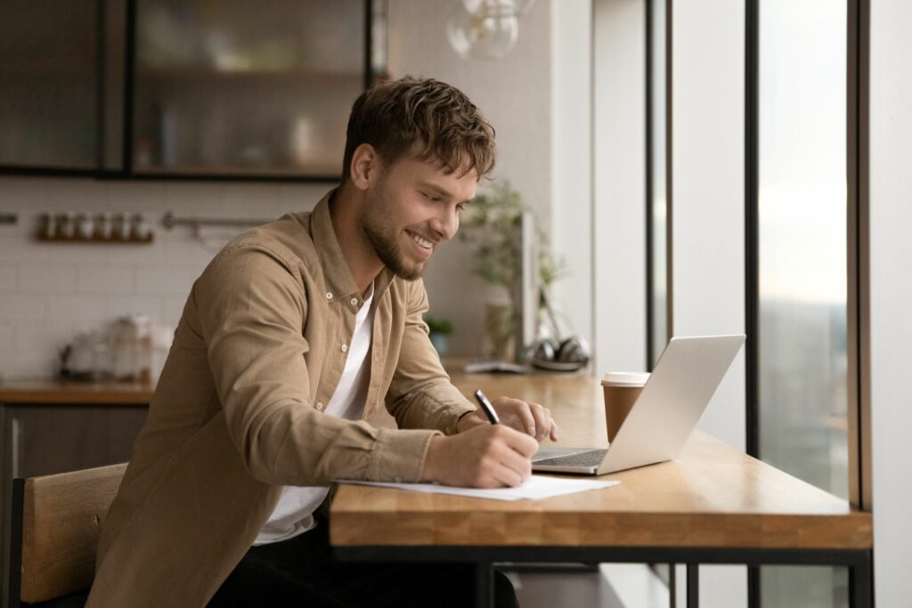 Australian man smiling and sitting at table looking at laptop researching to find a reliable tradesman. Person is holding a pen taking notes on the best tips to choosing a tradie.