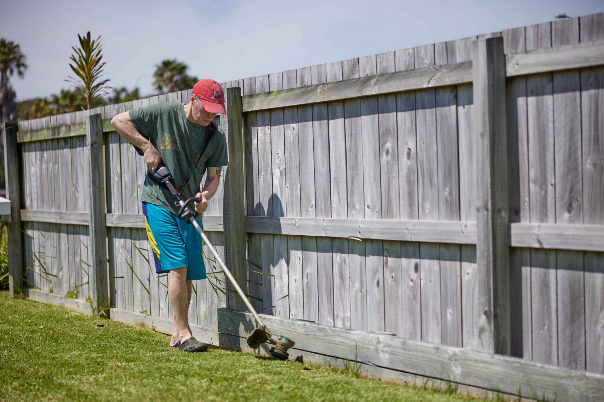 Man using whipper snipper against timber fence. Treated pine fence plinth is placed at the bottom of the fence palings to create a smooth edge to cut the grass against without damaging the fence